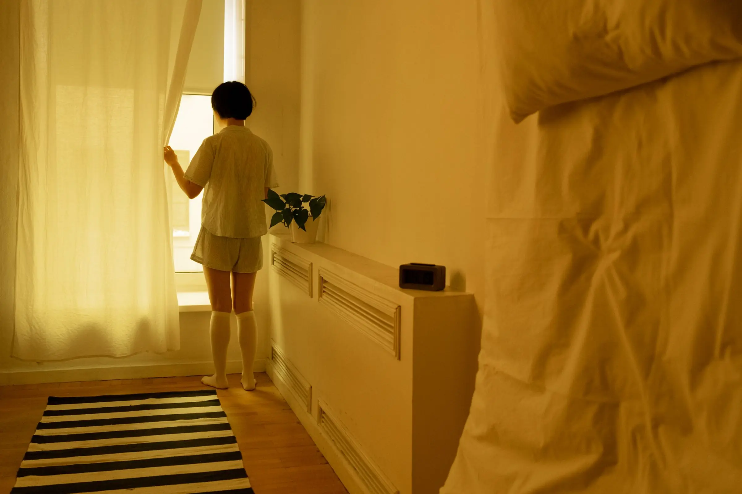 A woman in pyjamas pulling a curtain back on an ordinary morning, alone in a warm, sunlit room.